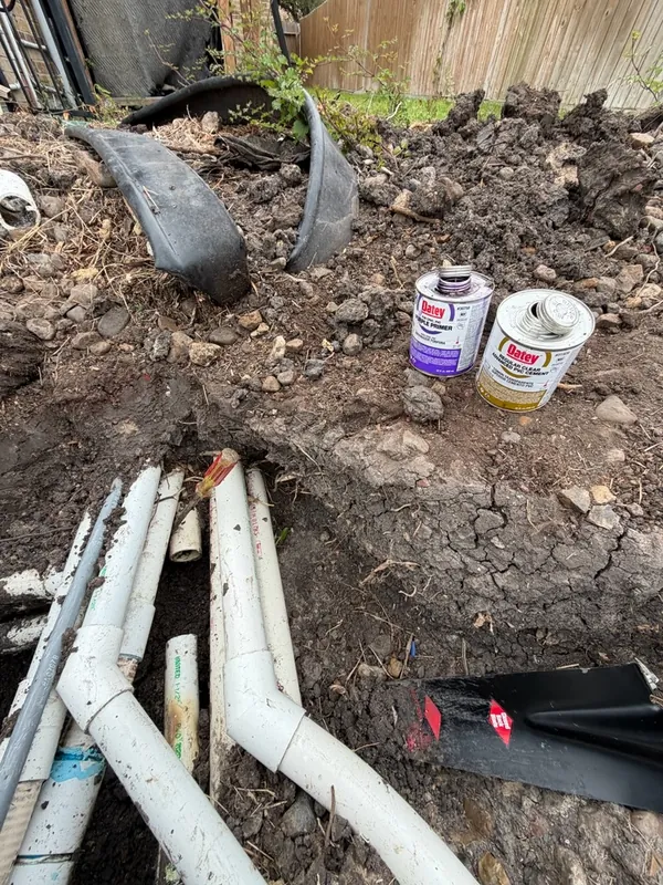 Exposed PVC drain lines in an open trench with primer and cement cans during a repair